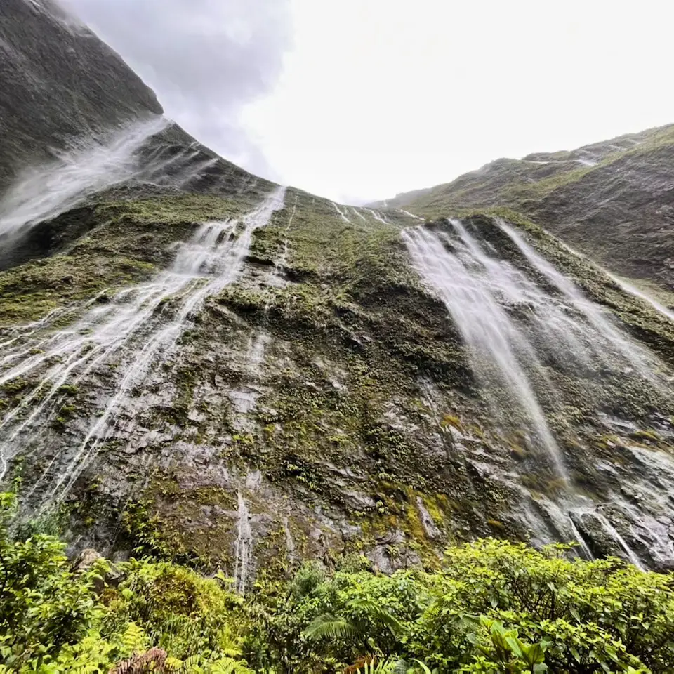 Several waterfalls pouring down a steep green cliff under cloud