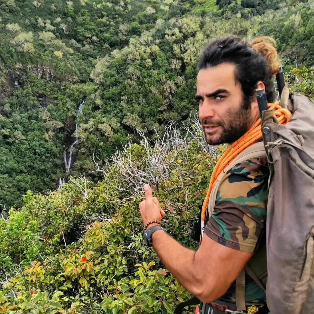 Renan Gobbet Hernandez with a rope pack on a jungle ridge, pointing toward a waterfall